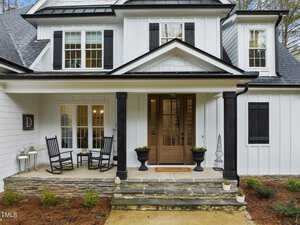 A modern white house with black shutters and roof, featuring a cozy porch with two rocking chairs and a small table. The wooden front door has glass panels, and the porch is decorated with plants and lanterns.