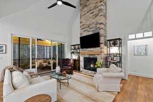 A cozy living room with a high ceiling features a tall stone fireplace with a TV above it. There are two cushioned chairs, a white couch, and a glass coffee table. Large windows lead to a screened porch with outdoor seating. Warm lighting fills the space.