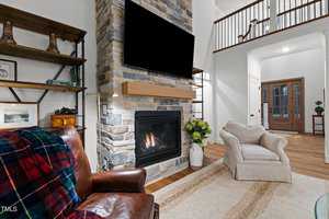 A cozy living room featuring a leather armchair with a plaid throw, a stone fireplace with a mounted TV, and a checkered armchair. Shelves display decor pieces. Natural light enters through the front door and windows, highlighting the hardwood floor.