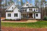 A white two-story house with a dark roof and black shutters, nestled in a wooded area of Wake Forest. It features a covered front porch with chairs, surrounded by neatly landscaped greenery and a curved walkway leading to the entrance at 3588 Cotton Field Court.