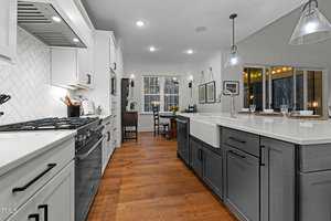 Modern kitchen with white and gray cabinets, hardwood floors, and recessed lighting. Features a large farmhouse sink, a gas stove with a white tile backsplash, and a dining area with large windows. Pendant lights hang over the island.