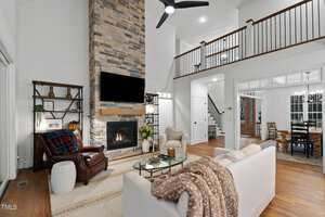 A cozy living room with a tall stone fireplace, a TV mounted above it, and a ceiling fan overhead. There's a sofa with a blanket, an armchair, and a small table. The room opens to a dining area with a large wooden table and chairs.