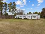 A white, single-story house with a metal roof sits serenely in the middle of a large, grassy field at 2910 Flat Rock Road, Franklinton. Surrounded by a few bushes and tall trees, the partly cloudy sky with patches of blue enhances this tranquil rural setting.
