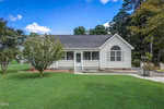 Nestled in the charming area of Franklinton, a small, single-story white house with a dark gray roof sits at 2512 Wes Sandling Road. Surrounded by verdant grass and trees, a concrete pathway guides visitors to its inviting screened front porch under a sky dotted with clouds.