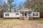 Located at 4129 Doe Court in Oxford, this white manufactured home boasts a brick foundation and a charming covered front porch. Surrounded by tall trees, it features black shutters on the windows and a neatly manicured lawn under a clear blue sky. Steps lead gracefully up to the inviting porch.
