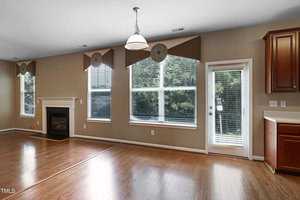 A spacious living room with wooden floors, featuring a fireplace, large windows with patterned shades, and a glass door leading outside. The walls are painted light brown, and a hanging light fixture is visible from the ceiling.