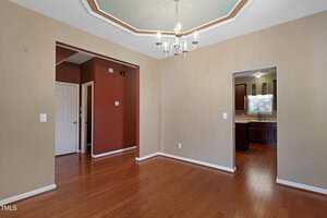 A dining room with hardwood floors features a chandelier hanging from a decorative ceiling. Walls are painted in beige and one deep red accent wall is visible. The room leads into a kitchen with dark wood cabinetry.