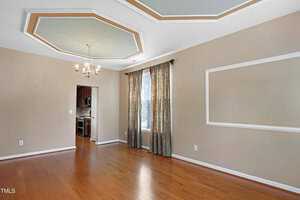 A spacious, empty dining room with hardwood floors, beige walls, and a decorative ceiling with two octagonal accents. A light fixture hangs above, and a window features floral curtains. An open doorway leads to a visible kitchen area.