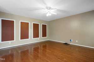 An empty room with wooden floors and beige walls features four brown rectangular panels framed in white. A ceiling fan is centered above, and there is a bundle of cables near one wall outlet.
