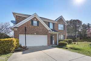 Two-story brick house with a double garage and a basketball hoop in the driveway. Surrounded by shrubs and trees under a clear blue sky.