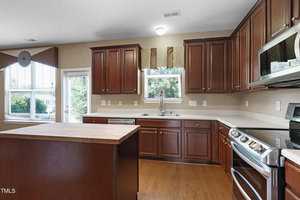 A modern kitchen with wooden cabinets, a central island, and stainless steel appliances. Natural light streams in through two windows with brown valances. The floor is wooden, complementing the cabinetry.