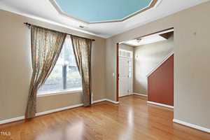A spacious room with hardwood floors, beige walls, and a window with patterned curtains. The ceiling has a unique octagonal design with a blue accent. A staircase is visible through an open entryway. Natural light brightens the space.