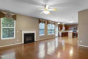 Spacious, empty living area with hardwood floors, beige walls, and a ceiling fan. The room features a white fireplace and large windows. An open kitchen with wooden cabinets and stainless steel appliances is visible in the background.