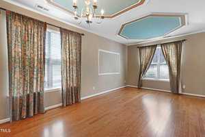 A spacious, empty room with hardwood floors and beige walls. Two large windows have floral patterned curtains. A chandelier hangs from a ceiling with teal and white geometric accents.
