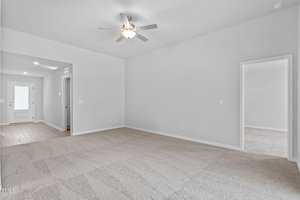 A bright, empty room with beige carpet and light gray walls. A modern ceiling fan is centered on the ceiling. Two open doorways lead to separate rooms, and a hallway leads to a door with a window, allowing natural light in.