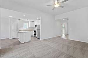 Modern, unfurnished open-concept living space with a view of the kitchen and living room. The kitchen features white cabinetry, stainless steel appliances, and a granite countertop island. A ceiling fan is installed in the living area with carpet flooring.