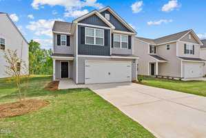 A two-story suburban house on 100 Scoville Street in Oxford boasts gray siding and white trim, complemented by a double garage and a neatly maintained lawn. The sky is blue with scattered clouds, and nearby houses are visible.