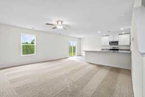 Spacious open-concept living area with beige carpet, light walls, ceiling fan, and recessed lighting. Features a modern kitchen with white cabinets, stainless steel appliances, and a granite countertop with bar seating. Large windows and glass doors offer natural light.