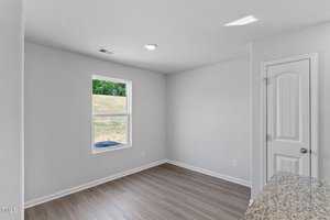 A small, unfurnished room with white walls and a single window letting in natural light. The floor is wood, and there is a closed white door on the right. A small section of a kitchen counter is visible in the bottom right corner.