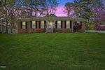 A single-story brick house at 104 Royall Rd, with white trim and black shutters, sits on a large Oxford lawn surrounded by trees. The sun sets majestically in the background, casting a pink and purple hue across the sky.