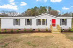 A white single-story house on 1635 Pope Road in Creedmoor features a red front door, black shutters, and a small wooden porch. The yard is landscaped with a line of small shrubs, and the roof is gray. Trees and a partly cloudy sky are visible in the background.