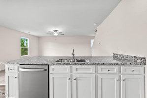 Modern kitchen with a large island featuring a granite countertop and built-in sink. The cabinets are white, and there is a stainless steel dishwasher. In the background, the living area is visible with carpeted flooring and a ceiling fan.