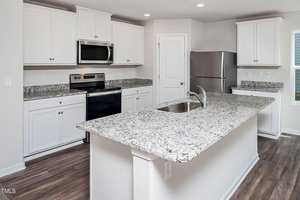 A modern kitchen with white cabinets, granite countertops, and stainless steel appliances, including a refrigerator, oven, and microwave. The kitchen features a large island with a sink and dark wooden flooring.