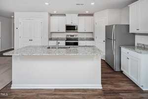 Modern kitchen with white cabinets, granite countertops, and stainless steel appliances, including a refrigerator, oven, and microwave. A large island with a sink is centered on a wooden floor. White walls and a double-door pantry are visible.