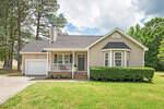 Located at 1090 Blackstone Drive in Creedmoor, this charming single-story house boasts beige siding, a black roof, and a green front door. A white railing porch and brick steps complete the look, with a single-car garage. It's surrounded by well-maintained shrubs and a lush green lawn against a backdrop of trees.