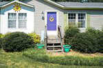 A charming small house on 552 Locks Lane features light gray siding and a distinctive purple front door with a sun ornament above its window. Two green planters grace the steps, displaying its number proudly. Nestled in Rougemont, the house is surrounded by lush bushes and greenery.
