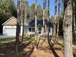 Nestled at 2106 Bedell Lane in Oxford, this small white single-story house with a dark roof is surrounded by tall pine trees. It boasts a charming front porch and a two-car garage. Sunlight filters through the trees, casting gentle shadows on the driveway and lawn.