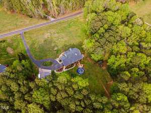 Aerial view of a house surrounded by dense green forest, with a curved driveway leading to it. There's a small pool in the backyard and a road visible nearby. The house has a spacious lawn and a wraparound deck.