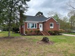 The red brick house at 204 Prospect Avenue, with its triangular roof, sits on a spacious grassy lawn in Oxford. A tree stump graces the foreground, while trees with budding leaves form the background. A small staircase with a railing leads to the front door beneath an overcast sky.
