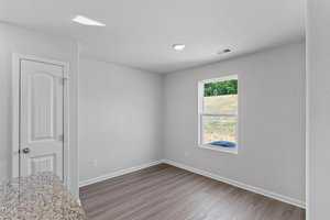 An empty room with light gray walls, a window on the right letting in natural light, and wood-like flooring. A door with a round knob is on the left. A speckled countertop is partially visible in the foreground.
