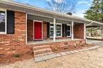 A charming brick house located at 617 29th Street boasts a covered front porch with white columns and a striking red door. The yard features a pathway of square stones leading to the steps, framed by plants along the foundation. A tree stands tall in the background beneath a vibrant blue sky.