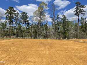 A cleared plot of land with brown soil, nestled at 206 Shelfit Trail, is surrounded by a line of tall trees under the Oxford blue sky with scattered clouds.