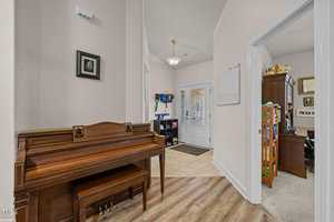 A cozy hallway with a wooden piano on the left, a door with a glass panel ahead, and a glimpse of an office on the right. The hallway features light-colored walls, wooden flooring, and a small chandelier.