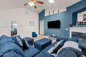 A cozy living room with a large blue sectional sofa facing a flat-screen TV mounted on a teal accent wall. There's a ceiling fan above, a fireplace on the right, and an open doorway leading to a well-lit room. Natural light fills the space.