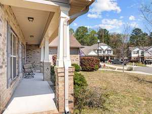Front porch of a suburban home with light brown brick, featuring a small seating area. The view includes a grassy yard and neighboring houses on a sunny day with a few clouds in the sky.