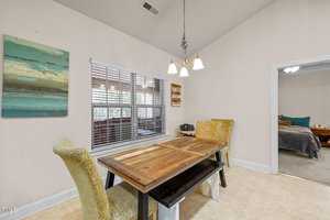 A dining area with a wooden table and two cushioned chairs. A painting hangs on the light-colored wall. A window with blinds is to the left, and a doorway leads to a bedroom with a bed and nightstand visible. A ceiling light fixture is above.