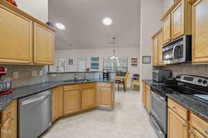 Kitchen with wooden cabinets, granite countertops, and stainless steel appliances including a refrigerator, microwave, and oven. A red stand mixer is on the counter. The kitchen leads to a bright dining area with a round table and chairs.