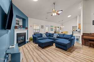 A spacious living room with a blue accent wall, featuring a large blue sectional sofa. There's a wall-mounted TV, a fireplace, and a ceiling fan. The room has wooden flooring and large windows letting in natural light. A piano is visible to the right.