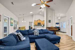 A spacious living room featuring a large blue sectional sofa with decorative pillows. The space is open to a modern kitchen with wooden cabinets. A ceiling fan hangs above, and there's a small piano near the entrance visible in the background.