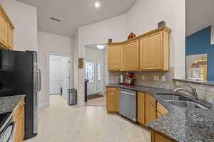 A bright, modern kitchen featuring granite countertops, light wood cabinets, stainless steel appliances, and a double sink. A red mixer sits on the counter. The kitchen opens to an entryway with a decorative glass door and a tiled floor.