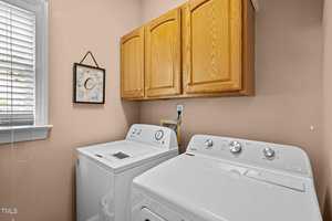 Laundry room with a washing machine and dryer beneath wooden cabinets. A framed picture hangs on the beige wall next to a window with blinds.