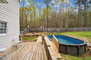 A backyard features a partially in-ground oval pool with a wooden deck and railing. Two white chairs sit on the deck, and a wooden privacy fence encloses the yard. Tall trees are visible beyond the fence under a blue sky with clouds.