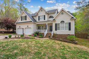 Nestled on 3711 Ben Mil Court in Franklinton, this charming two-story house features beige siding and a dark gray roof. The inviting porch with white railings and hanging plants complements the landscaped yard of grass, shrubs, and trees under a picturesque blue sky.