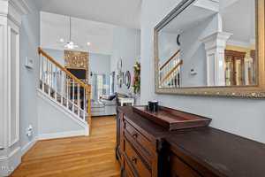 Hallway view of a cozy living room with wooden flooring. A large mirror hangs above a wooden dresser. Stairs lead up to a sitting area with a stone fireplace, TV, and sofa. The room is painted in soft blue tones with natural light streaming in.