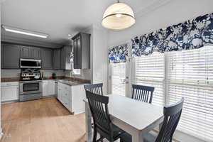 A kitchen with dark cabinetry, stainless steel appliances, and wood flooring. A dining area features a white table with dark chairs, framed by windows with floral curtains. A hanging light fixture illuminates the room.