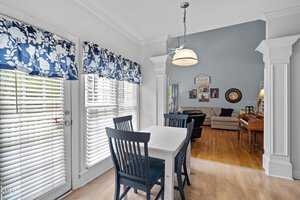 A dining area with a white table and two dark blue chairs is adjacent to a living room. The space features large windows with blue floral curtains, a hanging light fixture, hardwood floors, and a gray sofa with wall decor in the background.